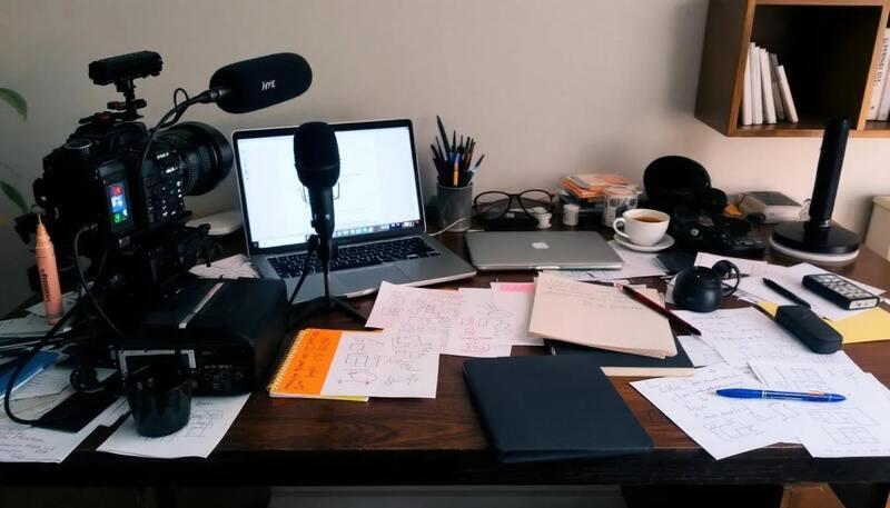 L'image montre un bureau désordonné avec divers équipements de médias, notamment une caméra, un microphone, un ordinateur portable et des notes éparpillées. Cet environnement évoque le quotidien d'un journaliste, où se mêlent recherche, rédaction et préparation d'articles pour la presse ou la télévision.